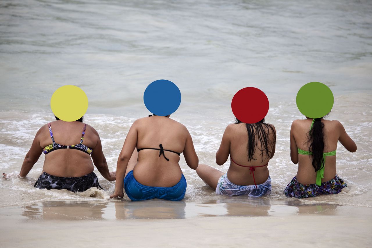 Foto de uma praia num dia cinza. Na beira do mar, 4 mulheres de roupa de banho sentadas de frente para o mar e de costas para o fotógrafo. Tapando a parte de trás de suas cabeças, cada mulher tem um círculo de cor diferente: amarelo, azul, vermelho e verde