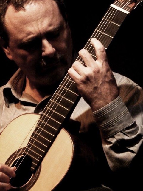 fundo escuro. Homem branco de camisa social clara toca um violão, que aparece parcialmente. O homem olha para sua mão esquerda tocando as cordas do violão.