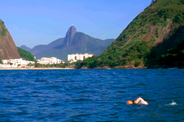 Foto da Baía da Guanabara com a Urca e o Pão de Açúcar ao fundo. Em primeiro plano, um homem nada no mar azul. 
