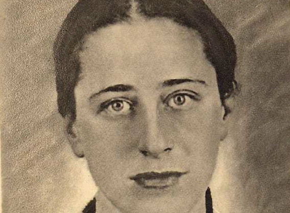 Foto em preto e branco, fundo cinza. Ao centro, o rosto de Olga Benário. Mulher branca, cabelos pretos presos, olhos claros.