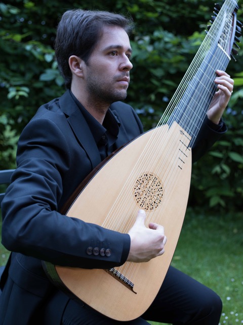 Foto do violonista Vinicius Perez tocando um alaúde barroco. O violonista é um homem branco, de cabelos pretos curtos. Usa camisa social preta de mangas longas e calça escura. Ele está sentado em um jardim, com folhagens ao fundo.