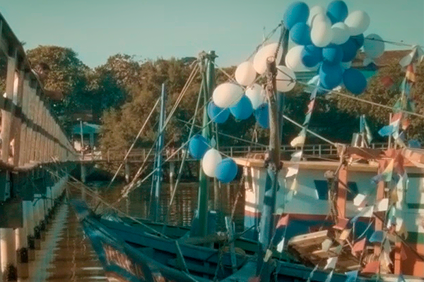Foto de um cais. Dia de sol, céu azul, árvores ao fundo. Ao lado esquerdo, a estrutura de uma ponte. Ao lado direito, um barco, em cujos mastros foram pendurados balões azuis e brancos. 