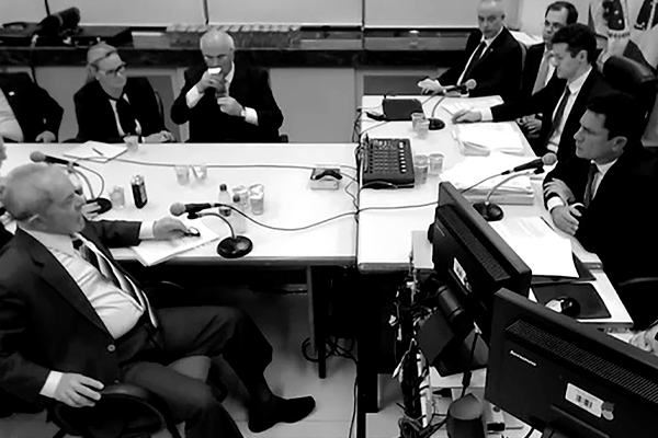 A foto, em preto e branco, mostra uma sala de reunião com uma mesa em formato de ‘T’, com computadores, microfones, copos e água. Há sete homens e uma mulher, todos brancos. Eles estão sentados, usam paletó e gravata. Ela usa terninho e blusa com laço na frente. Um dos homens (ex-juiz Sérgio Moro) indaga o outro (atual presidente, Luiz Inácio Lula da Silva) no que parece ser uma audiência da Operação Lava a Jato.