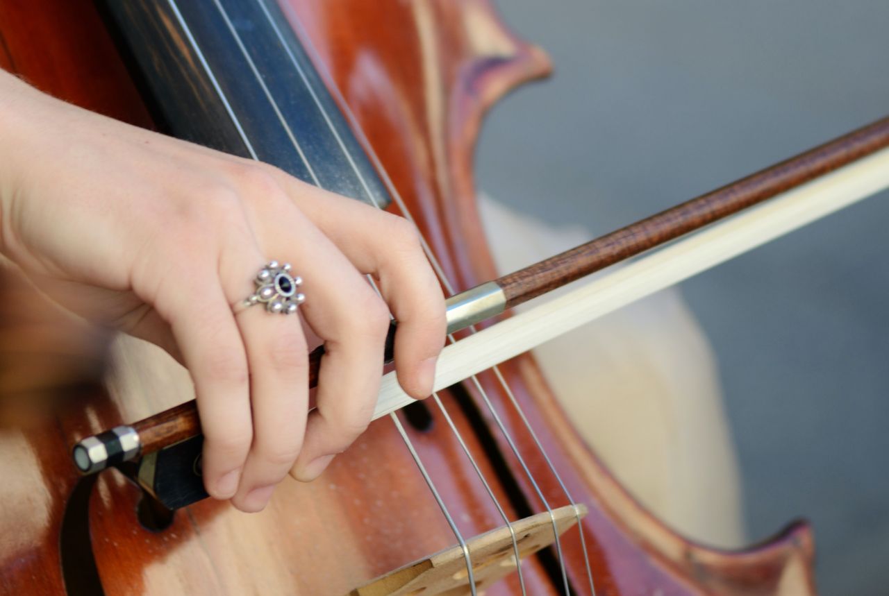 Foto da mão esquerda de uma mulher tocando um violoncelo. A mão é branca e tem um anel com pedras pequenas no anelar. Ela segura o arco do instrumento, do qual aparecem apenas parte do corpo e as cordas.