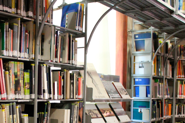 Foto de uma parte da biblioteca do CCJF. Do lado esquerdo e direito, estantes de ferro com as prateleiras repletas de livros. Entre elas, uma bancada com revistas.