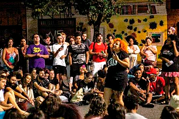 Foto de um evento Slam, batalha de poesias. Vários jovens sentados ao chão e outros em pé, todos em volta de uma jovem branca, cabelos cor de cenoura, bermuda e camiseta pretos, que declama para a platéia.