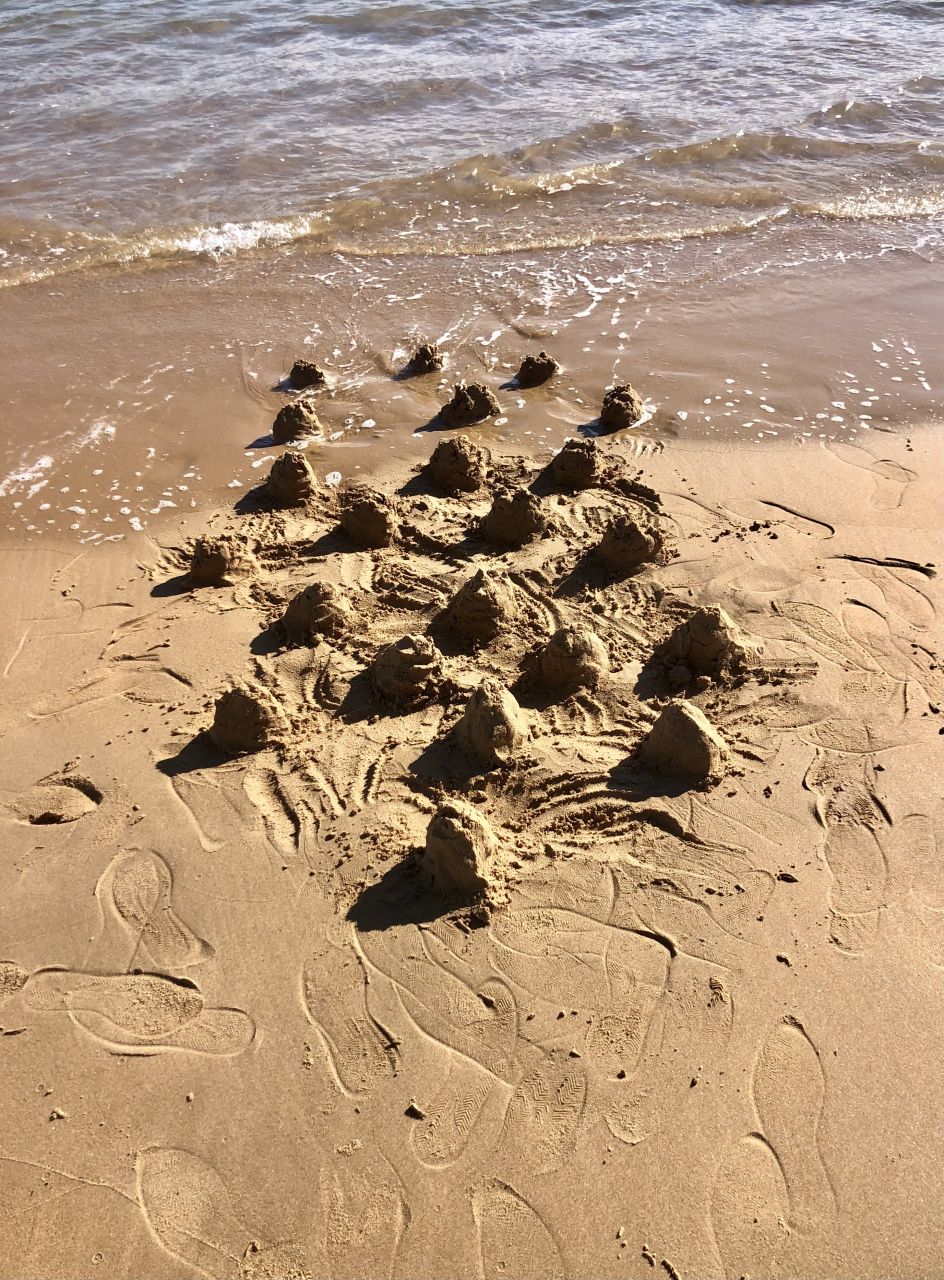 Foto. À beira-mar, uma série de montículos de areia entre a praia e a orla marítima. Alguns dos montículos já estão sendo desfeitos pelo mar. Entre os montículos, o artista riscou a areia, ligando-os uns aos outros. Ao redor, marcas de pegadas humanas, calçadas por sapatos, em várias direções.