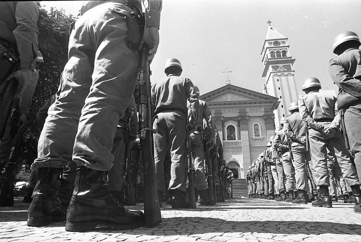 Na foto em preto e branco, alguns soldados aparece de costas, fardados, em fila de frente para uma igreja com construção histórica. 