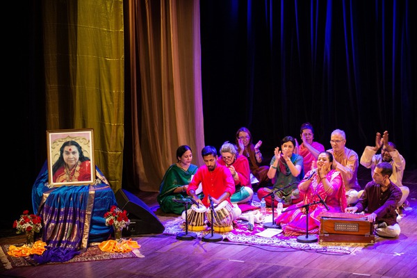 Na foto, um grupo de pessoas, entre homens e mulheres, estão sentadas em um palco cantando e batendo palmas. Elas estão sentadas de perna cruzadas. Vestem trajes indianos coloridos. No primeiro plano, dois homens tocam instrumentos musicais. A luz que ilumina o palco é azulada. No canto esquerdo, uma cadeira com tecidos azuis com detalhes dourados repousa uma foto da mestra indiana Shri Mataji Nirmala Devi. No plano de fundo, cortinas de veludo cobre, dourada e azul compõem o cenário. 