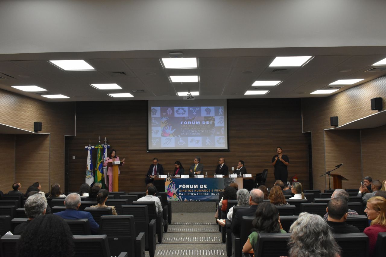 Na foto, o auditório do TRF2 e, ao fundo, uma gestora de comissão do fórum palestra em pé, ao lado da mesa do evento, que conta com cinco magistrados.